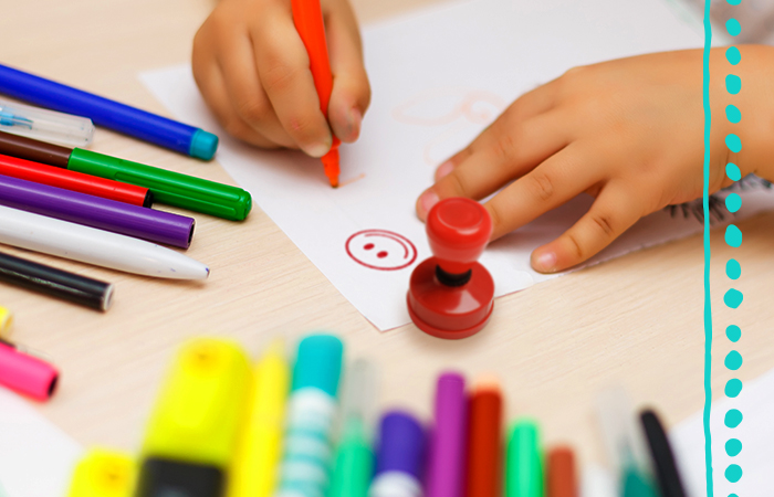 Hands drawing a red smiley face on a piece of paper surrounded by markers