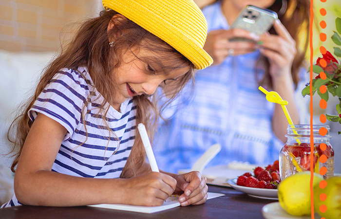 Child wearing a yellow hat sitting outside at a table writing in a book. 