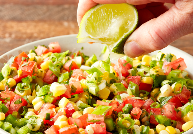 Corn, diced tomatoes, sliced green onion, minced jalapeno mixed in a bowl and a hand squeezing a lime over the bowl
