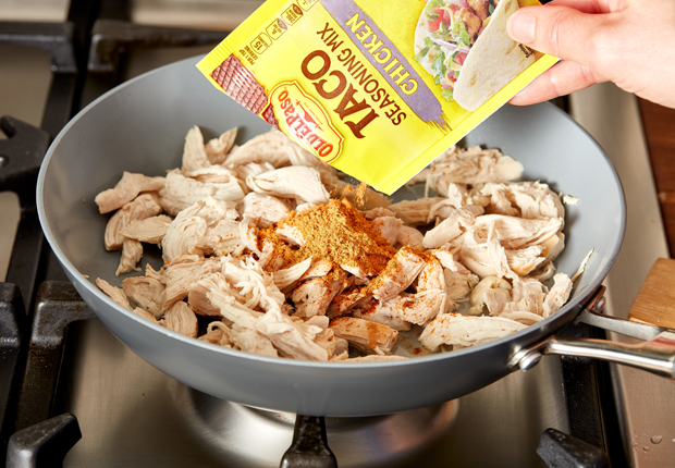 Shredded chicken in a skillet on the stove with a hand pouring a packet of Old El Paso Chicken Taco seasoning, front of package, over the chicken