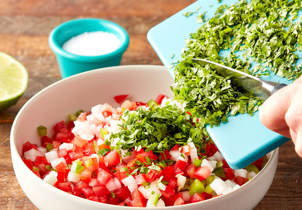 Diced tomato, diced white onion, minced jalapeño in a bowl and a cutting board above the bowl with chopped cilantro with a hand using a knife to scrape some cilantro into the bowl
