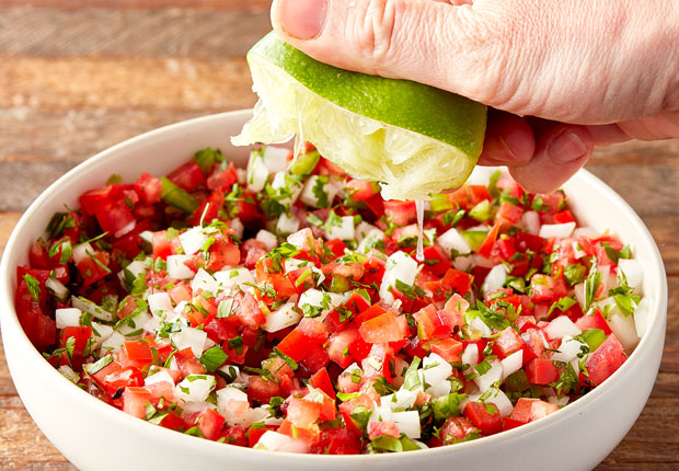 Diced tomato, diced white onion, minced jalapeño, chopped cilantro mixed in a bowl with a hand squeezing a lime above the bowl